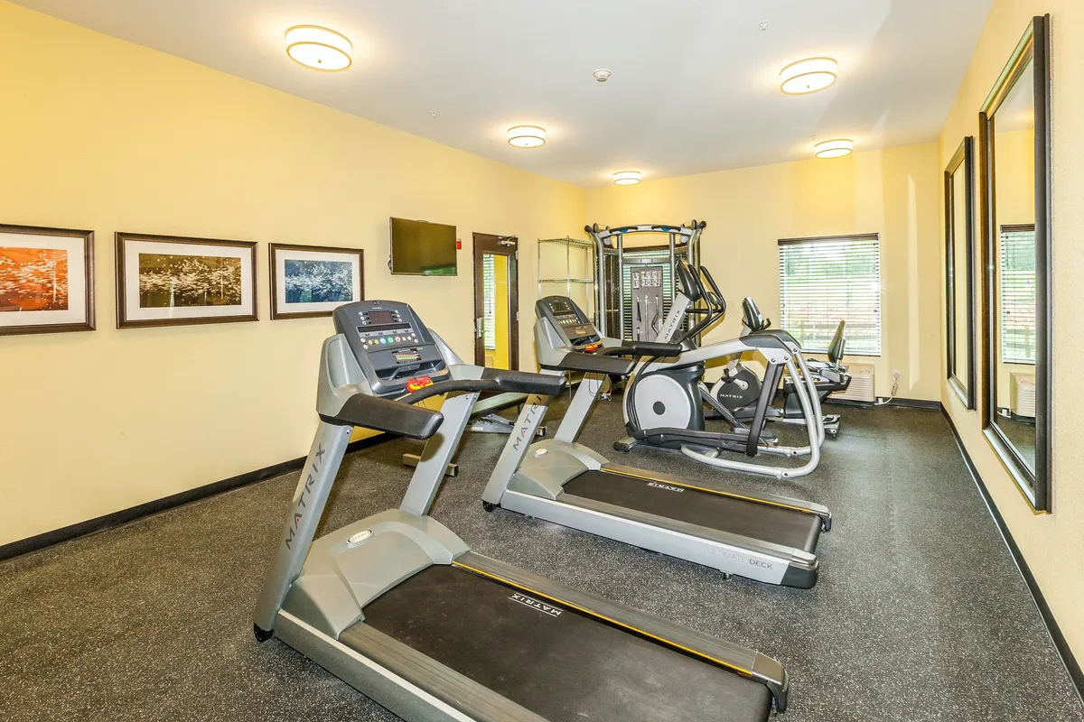 A well-equipped fitness center featuring treadmills, an elliptical machine, a weight machine, and large mirrors. Bright lighting and framed pictures contribute to the energetic and inviting environment.