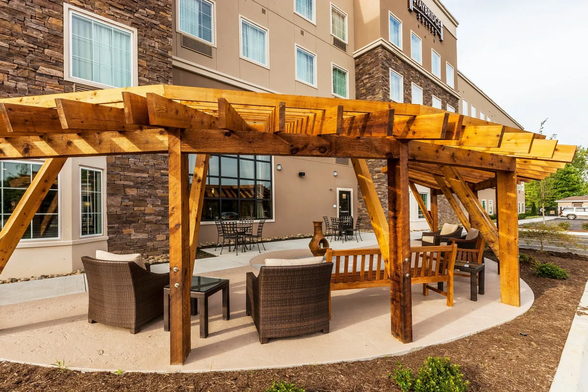 This image displays a wooden pergola providing shade over an inviting outdoor seating area framed by the stone exterior of a building. Groups of people are relaxing, enjoying coffee, and conversing, suggesting a social, leisurely atmosphere just outside the hotel.
