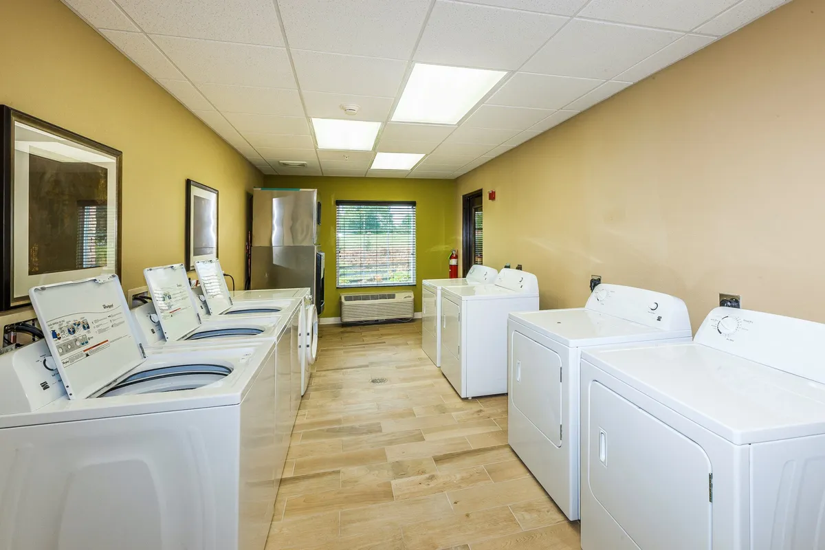 A practical laundry room with washing machines and dryers lined up by large windows. The scene suggests convenience for guests handling extended stays, with someone folding fresh laundry in a tidy, communal setting.
