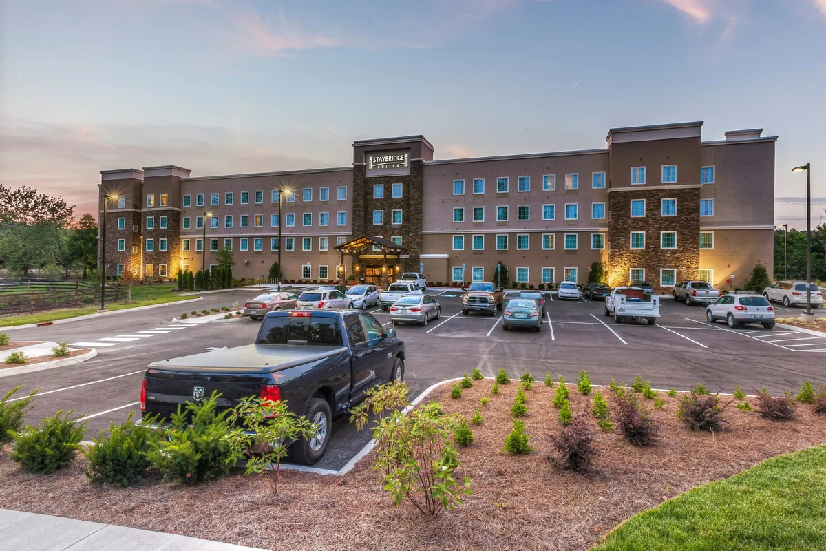 An inviting image focused on the hotel entrance, surrounded by a well-kept landscaped area and a parking lot. There are subtle hints of social interaction and the warmth of a welcoming arrival experience.