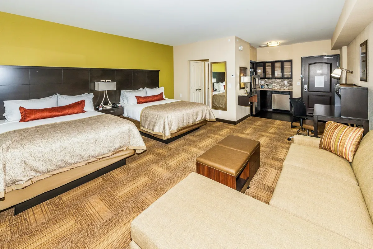 A well-lit studio interior featuring two queen beds side by side, a cozy sofa seating area, and a desk with a chair. The space is arranged to showcase a seamless transition between the sleeping, lounging, and working areas, with personal touches in decor and natural light streaming in.
