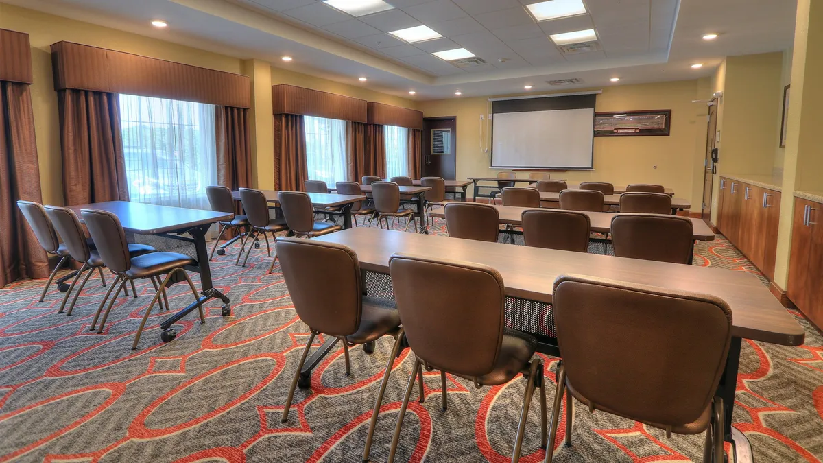 A modern meeting space featuring tables and chairs arranged for a team presentation, with a projector screen at the front and large windows letting in natural light. The setting is focused, professional, and conducive to collaborative work or business meetings.