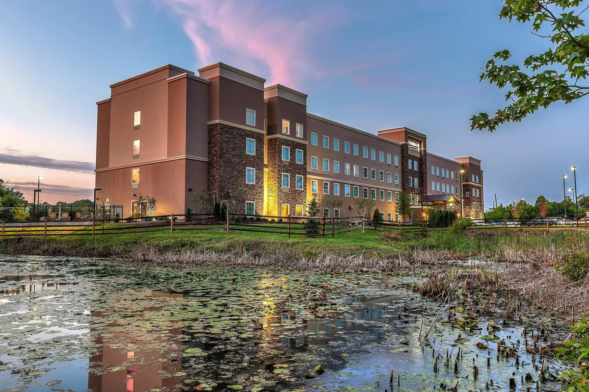 Overlooking a serene pond in the foreground, this exterior shot includes the hotel building and a sky tinged with the colors of sunset. The scene hints at possibilities for evening strolls, relaxation by the water, and outdoor gatherings.