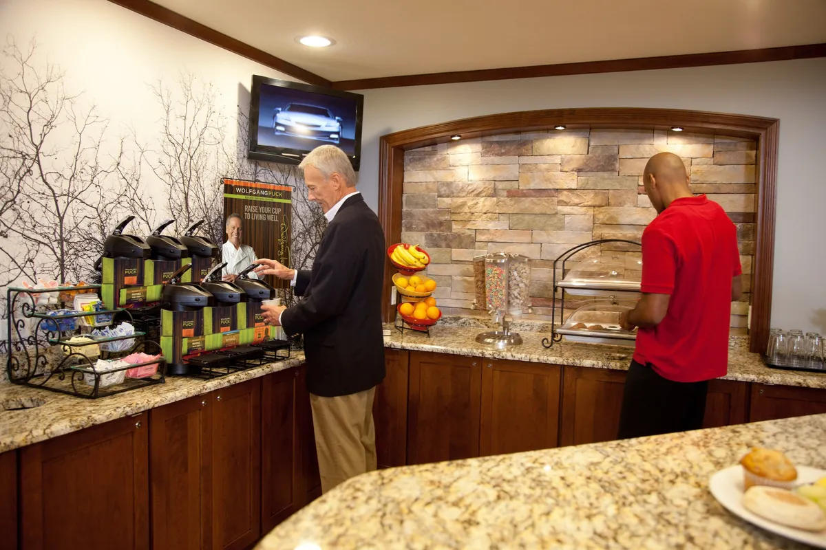 This image shows a welcoming interior breakfast area featuring coffee machines, a fruit display, a television, and a textured stone wall. The space is set up for informal dining, with elements like fresh fruit and hot beverages suggesting meal preparation and enjoyment.